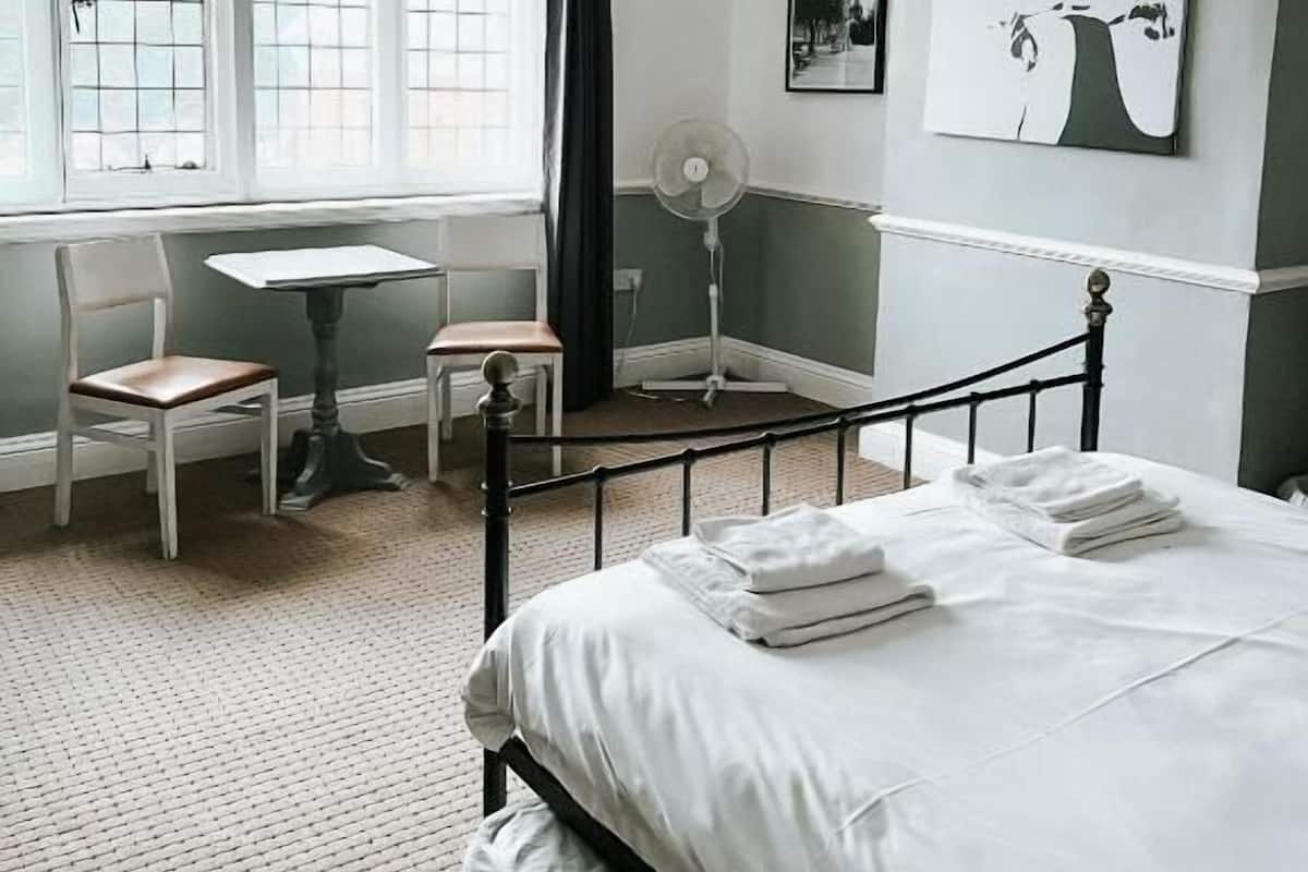 A bedroom with a bed, a desk, two chairs, a standing fan, and artwork on the walls.