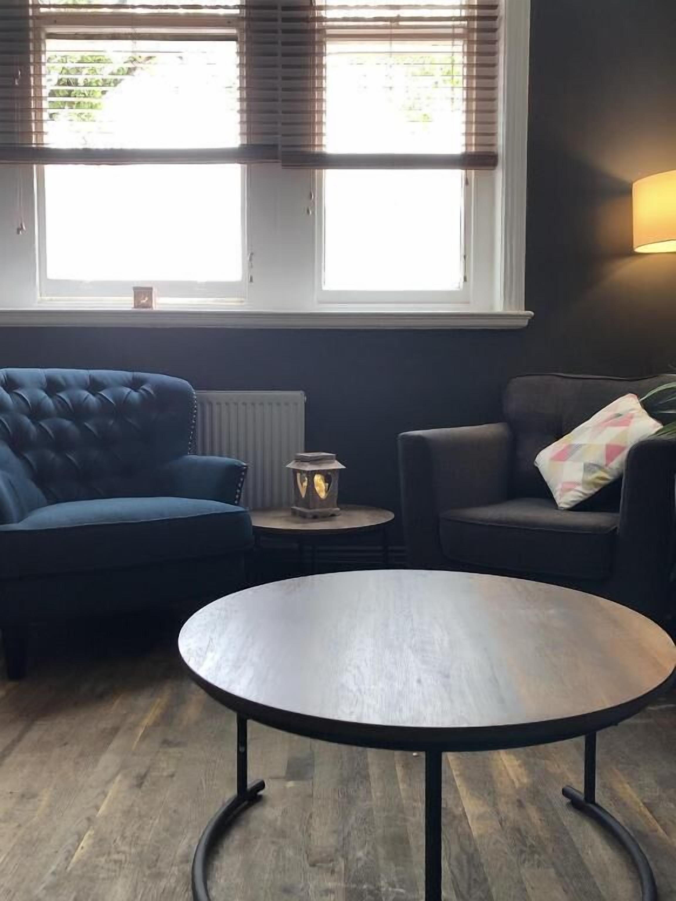Una habitación con dos sofás oscuros, una mesa redonda de madera, una mesita con un farolillo y una ventana con persianas.