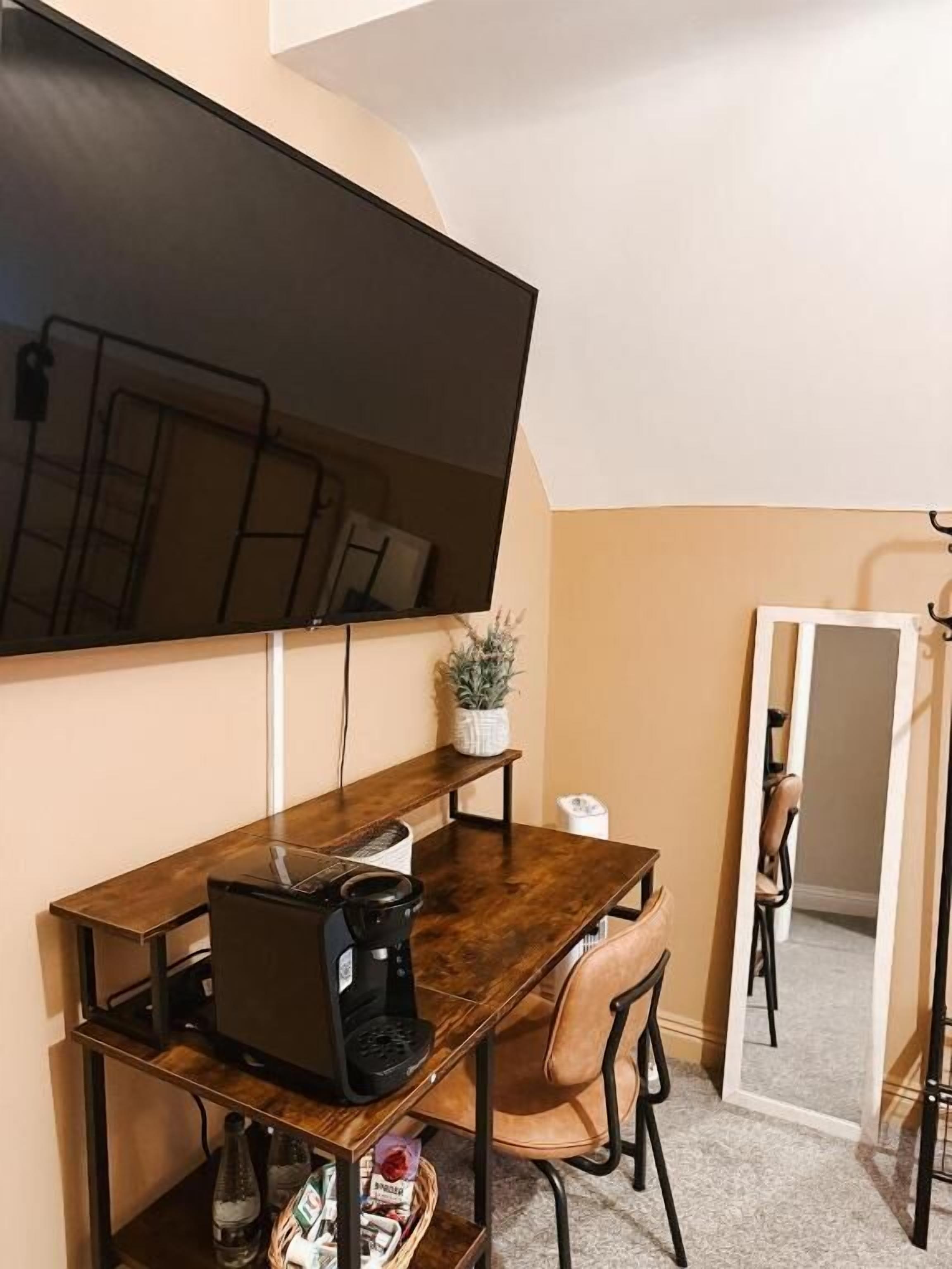 Een kleine, moderne kamer met een donker houten bureau, een zwarte koffiezetapparaat, een laptop en een enkele stoel. Een groot flatscreen-televisie is aan de muur boven het bureau bevestigd.