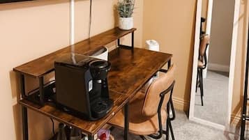 Een kleine, moderne kamer met een donker houten bureau, een zwarte koffiezetapparaat, een laptop en een enkele stoel. Een groot flatscreen-televisie is aan de muur boven het bureau bevestigd.