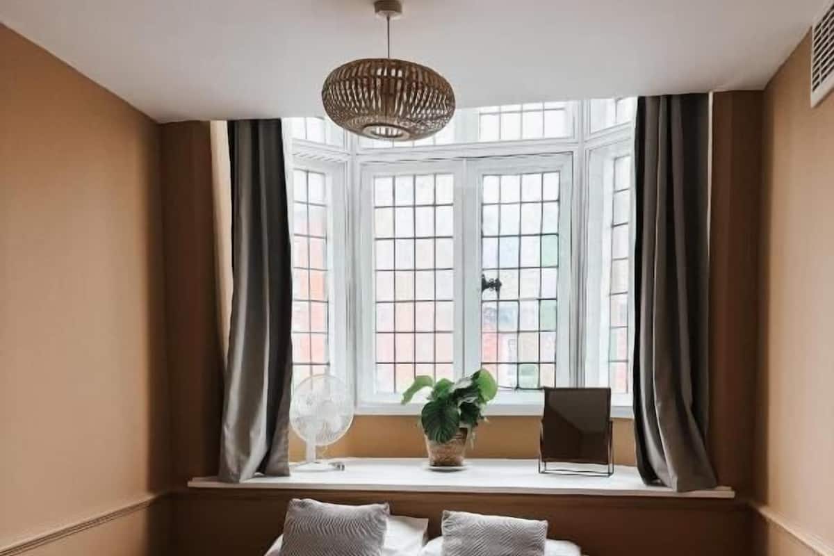 A bedroom with a large bed, a window with curtains, a plant on the windowsill, and a ceiling light fixture.