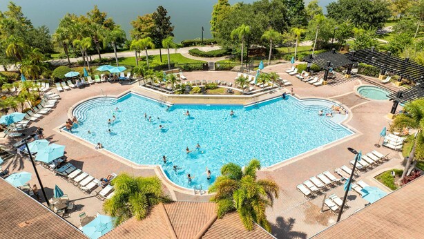 A large outdoor swimming pool surrounded by lounge chairs and umbrellas, with palm trees and a view of a lake. - Disney Getaway! Townhouse with Private Pool. Book Now! #VC304 (Orlando)