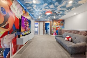 Une pièce décorée d’une fresque représentant Mario et Luigi, avec un canapé gris agrémenté d’un coussin à l’effigie de Mario, et un plafond orné de motifs de nuages.