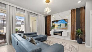 A modern living room with a sectional sofa, a flat-screen TV mounted on a wooden panel, and a large window with curtains.