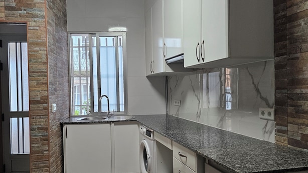 A modern kitchen with white cabinets, a granite countertop, a built-in washing machine, and a window with a view of the outside. - Real 43 *Good road (Neda)