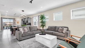 A modern living room with a grey sofa set, a white coffee table, and a green armchair. There is a dining area in the background with a table and chairs.