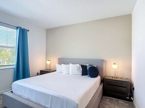 Una cama bien hecha con ropa blanca, cabecera gris y varios almohadones. Hay dos mesitas de noche con lámparas y una ventana con cortinas azules.