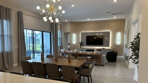 A modern living room with a dining area, a large flat-screen TV, and a large window with curtains. - Last Minute Disney! Private Pool, Game Room & Themed Bedrooms. Book Now! #144011 (Orlando)