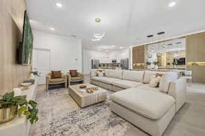 A modern living room with a sofa, chairs, a coffee table, and a kitchen in the background. - Last Minute Disney! Private Pool, Game Room & Themed Bedrooms. Book Now! #118059 (Orlando)