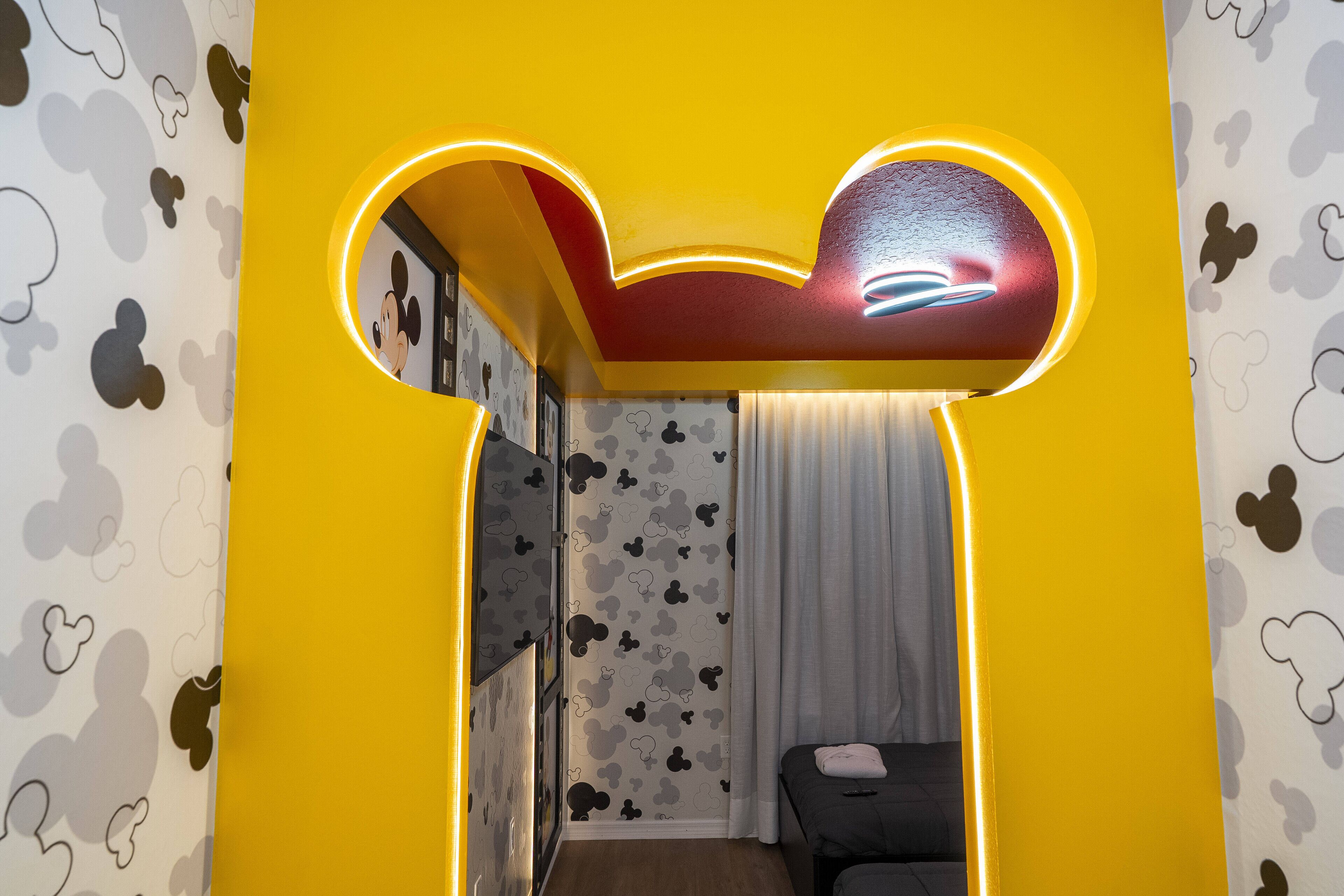 Una stanza con un soffitto a tema Topolino, un letto, una finestra con tende e una parete decorata con un motivo raffigurante Topolino.