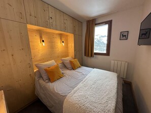 A bedroom with a wooden headboard, a bed with striped bedding and two yellow pillows, a window with curtains, and a radiator.