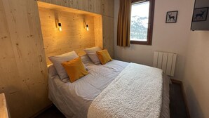 Une chambre à coucher avec une tête de lit en bois, un lit avec une literie à rayures et deux oreillers jaunes, une fenêtre avec des rideaux et un radiateur.