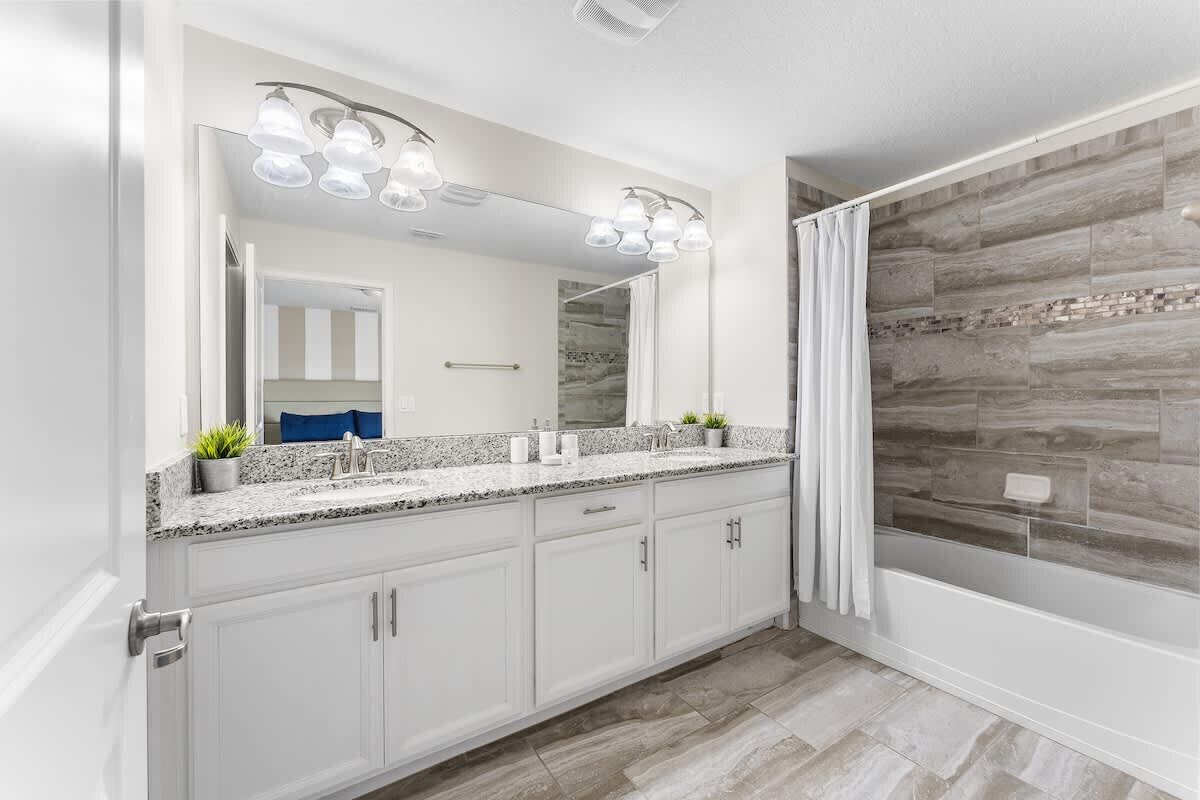 Une salle de bain moderne avec un comptoir en granit, des armoires blanches, un grand miroir et une douche dont le mur arrière est recouvert de pierres.
