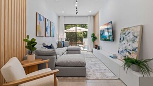 A modern living room with a grey sectional sofa, a wooden side table, and a potted plant. There are abstract paintings on the walls and a large window with an umbrella outside.