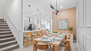 A modern dining area with a wooden table set for six, a staircase on the left, and a kitchen in the background.