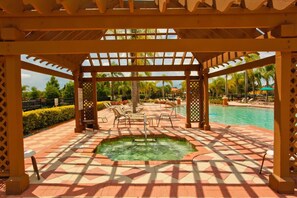 Ein sonniger Außenpoolbereich mit Whirlpool, Liegestühlen und einer Pergola.