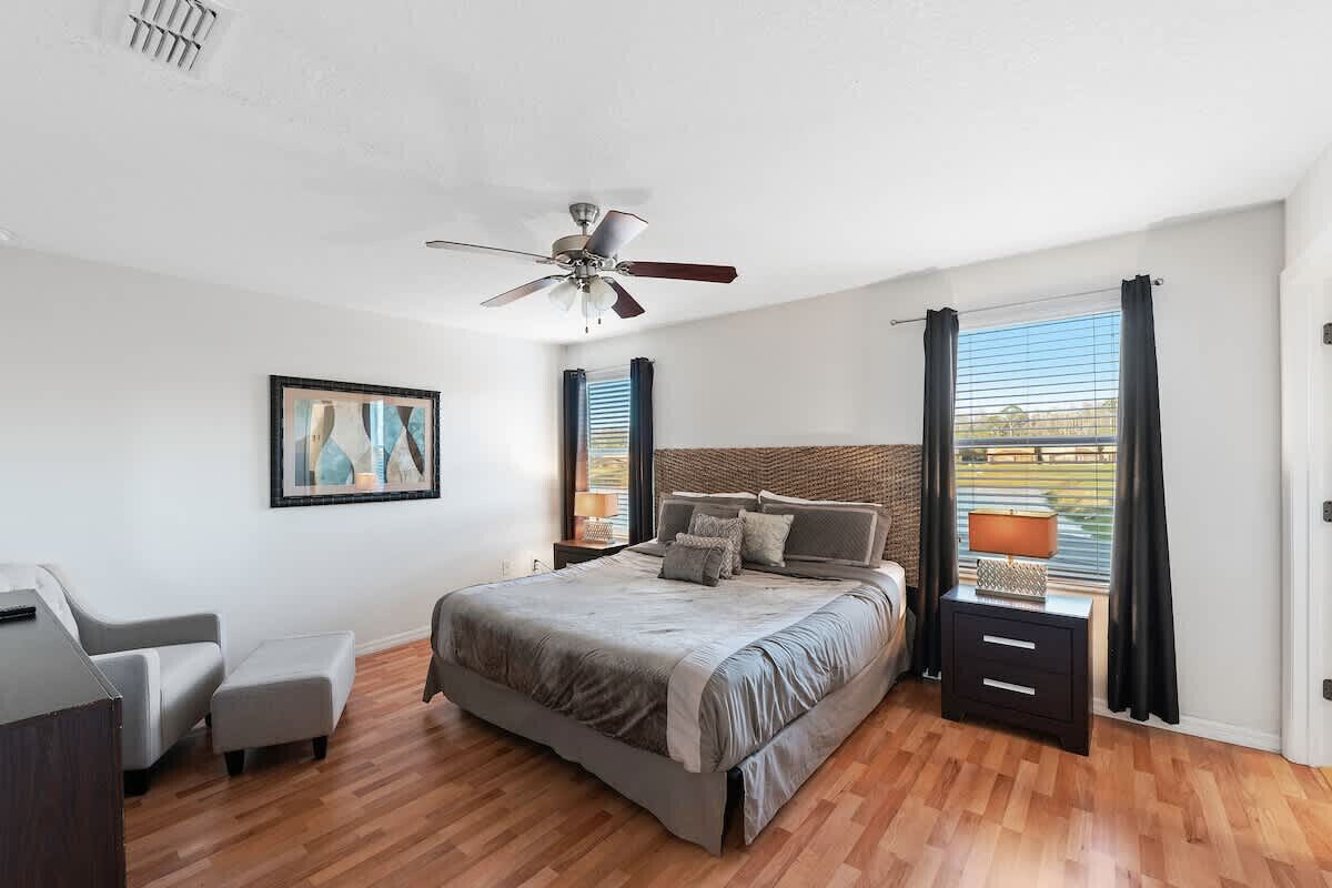 Une chambre à coucher comprenant un lit, un ventilateur de plafond, une table de chevet, une chaise et une fenêtre avec des rideaux.