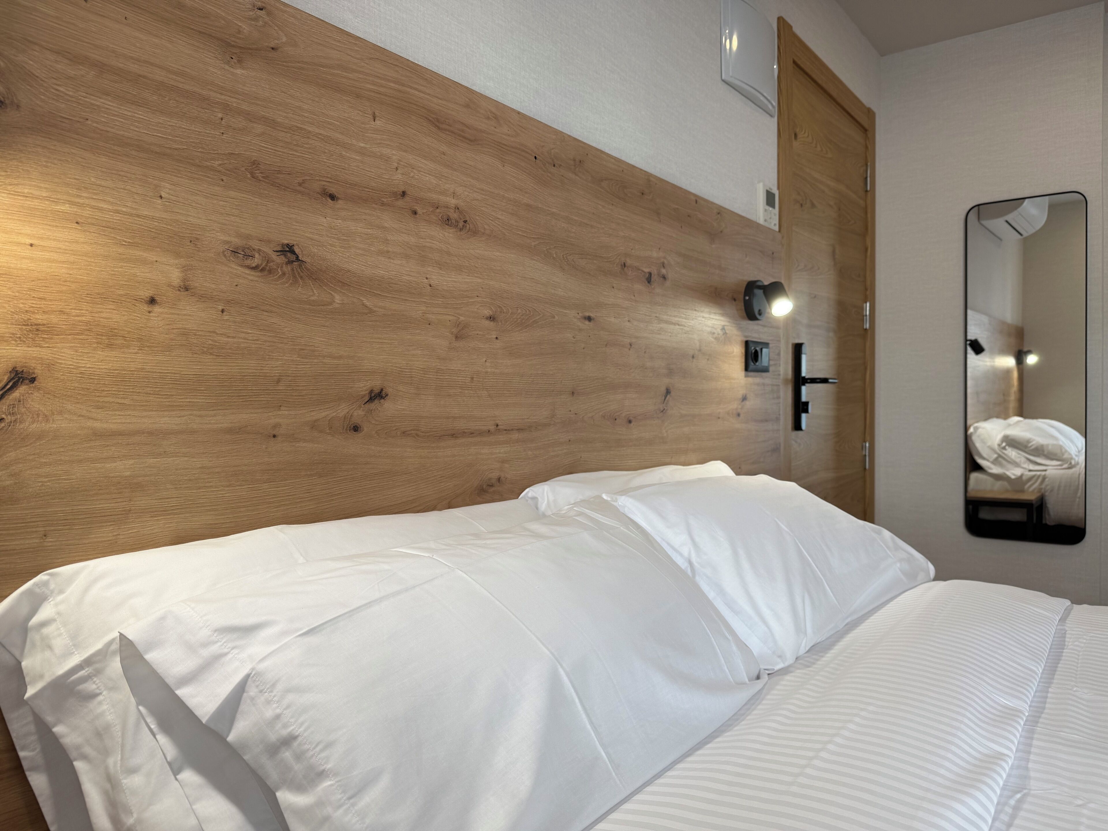 Hotellihuone, jossa on puinen sängynpääty, valkoisilla lakanoiden koristeltu sänky, yöpöytä lampun kanssa ja peili seinällä.