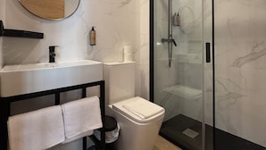 A modern bathroom with a marble wall, a round mirror, a white sink, a toilet, and a glass-enclosed shower.