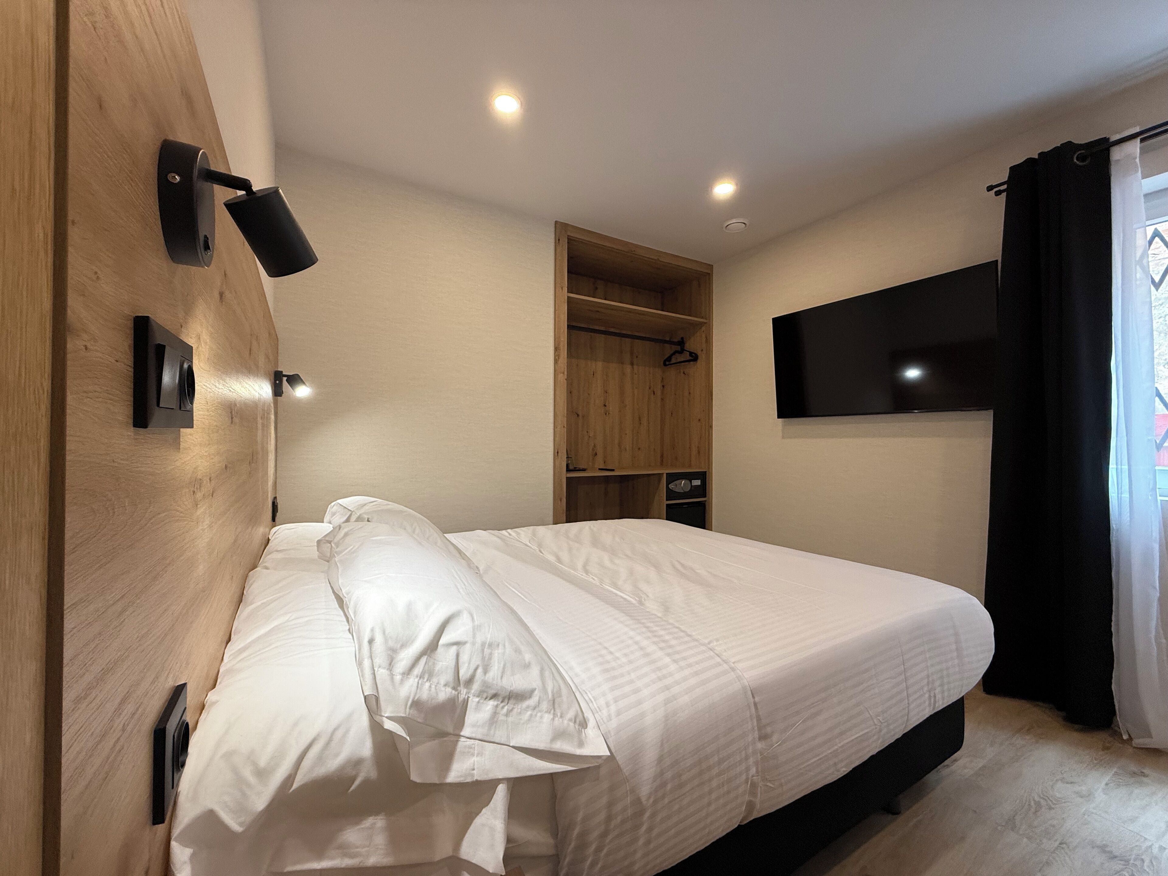 Una camera d'albergo moderna con un letto grande, una testiera in legno, un armadio a muro, una TV a schermo piatto montata sulla parete e una finestra con tende.