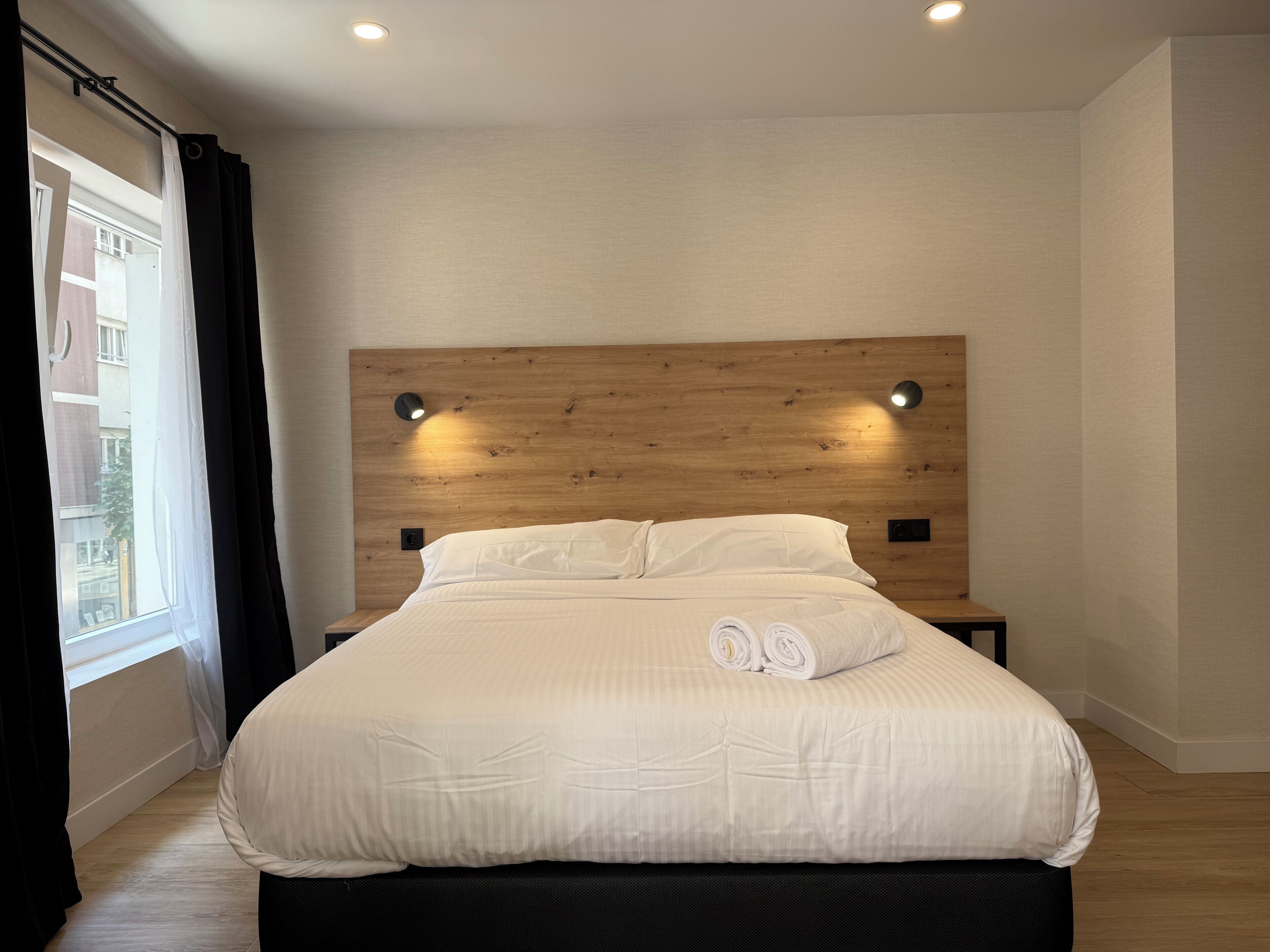 Un dormitorio moderno con cabecera de madera, dos lámparas de noche y una cama bien tendida con toallas enrolladas.