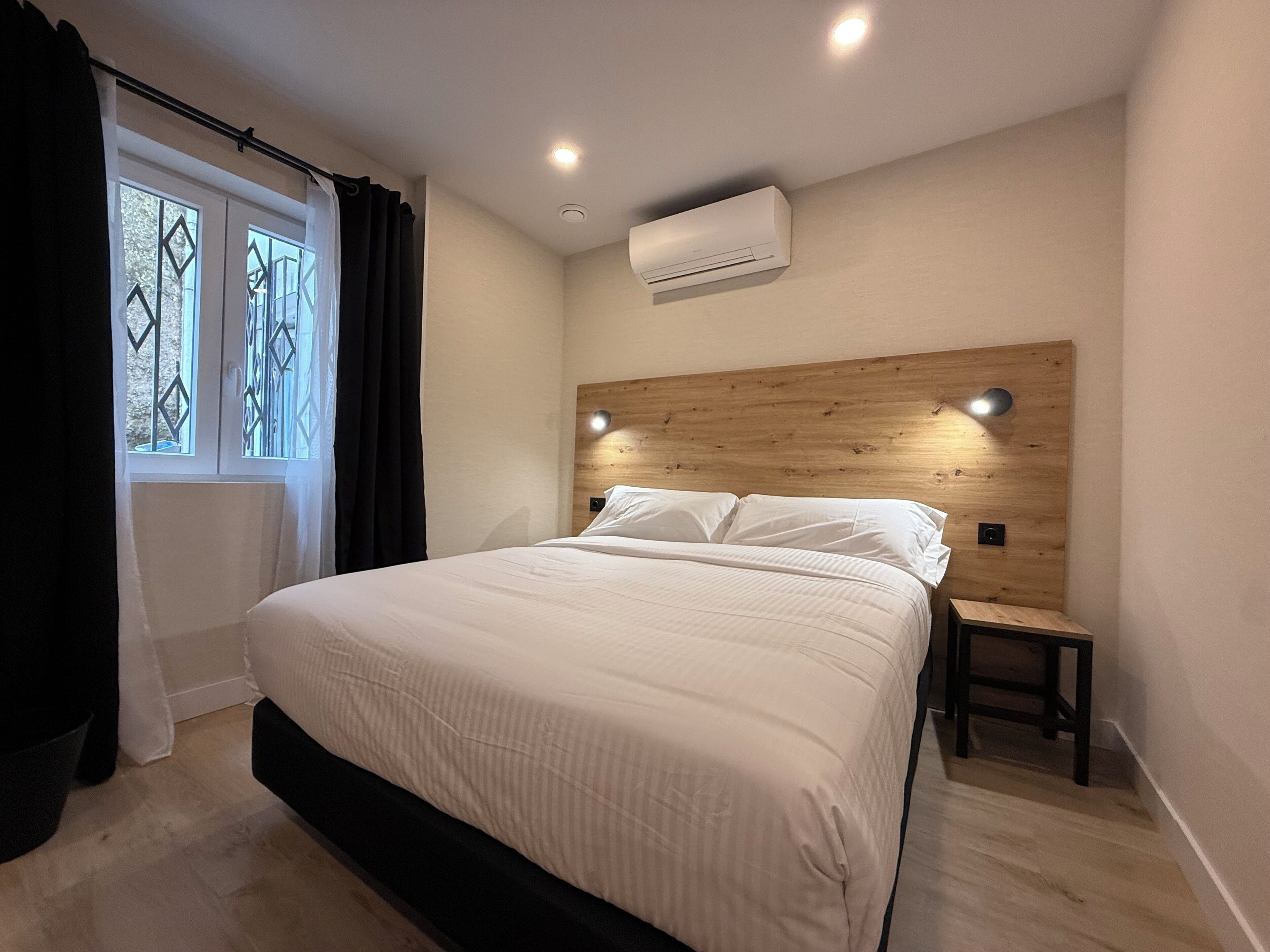 A bedroom with a bed, a nightstand, a window with curtains, and an air conditioning unit.