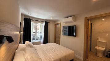 Un dormitorio con una cama, un televisor en la pared, un equipo de aire acondicionado y vistas al exterior a través de una ventana con cortinas.