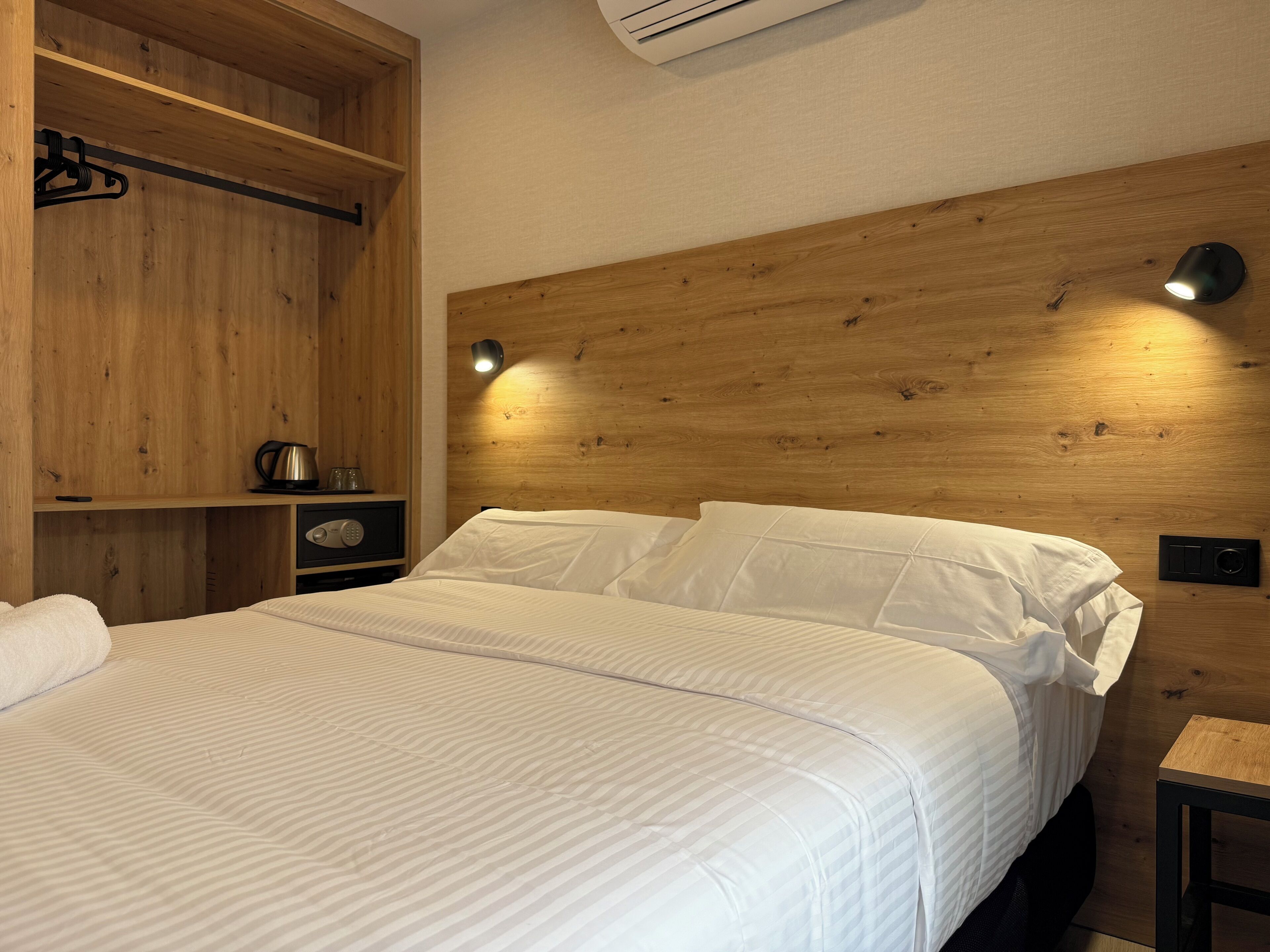 A neatly arranged bedroom with a wooden headboard, a bed with white linens, a bedside table with a lamp, and a shelf with a kettle and glasses.