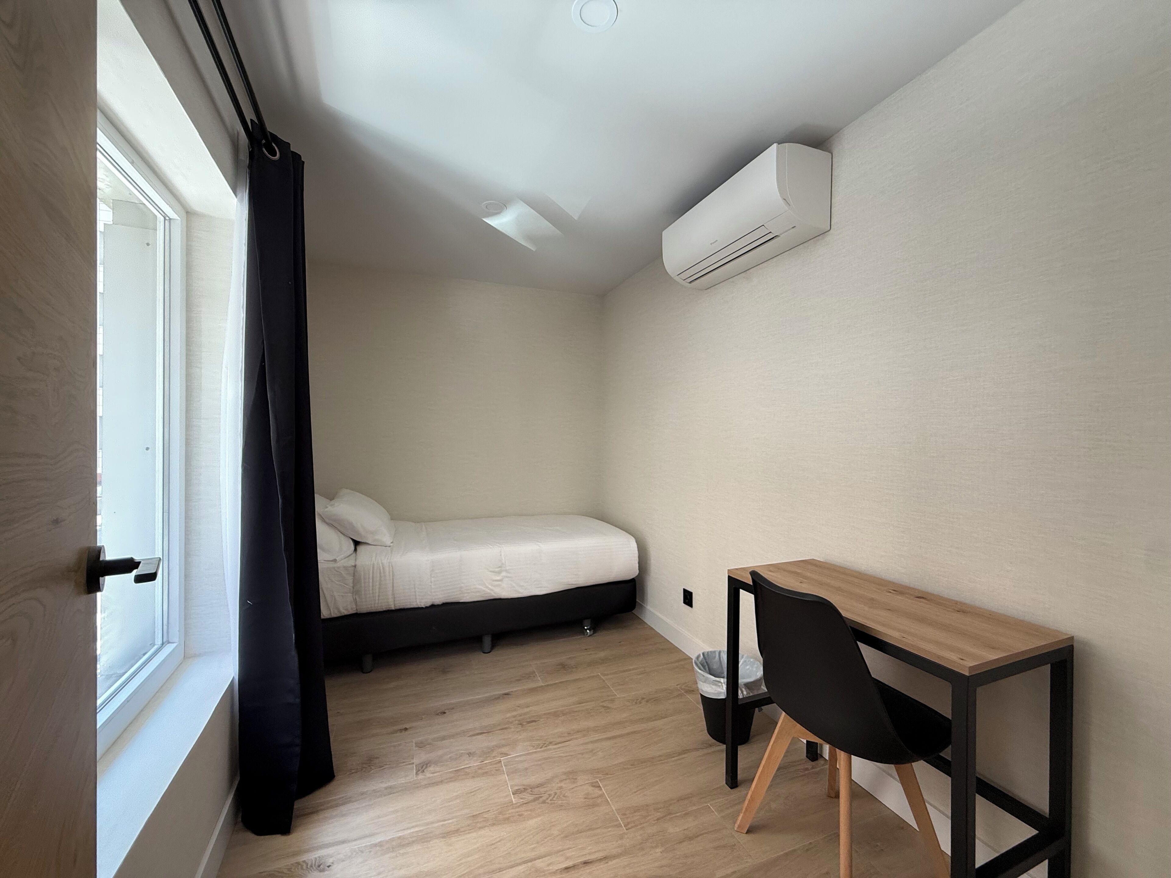 A small, modern room with a bed, a desk, and an air conditioning unit on the wall.