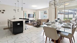 A modern kitchen with a central island, a dining area with a glass table, and a living space with a sofa and TV. - Disney Getaway! Private Pool & Themed Bedrooms. Book Now! #SLR506 (Orlando)