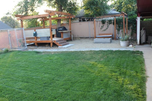 Un jardin avec une pergola en bois, un barbecue et un espace détente comprenant un banc et une table.