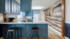 A kitchen with blue cabinets, a wooden island with stools, and a rustic wall with wooden planks. - The Nest with Firepit & Backyard Swings (Klamath Falls)