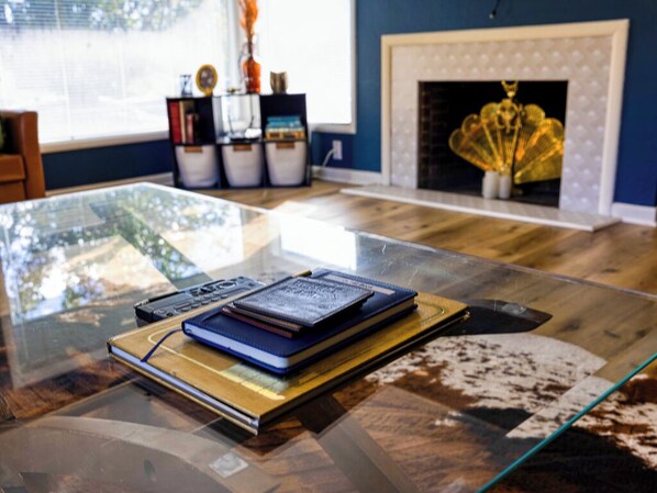 Una mesa con cubierta de vidrio, libros y una computadora portátil, una chimenea con un adorno, y una ventana con persianas.