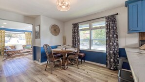 A dining area with a round wooden table and chairs, a ceiling light fixture, and a window with floral curtains. - The Nest with Firepit & Backyard Swings (Klamath Falls)