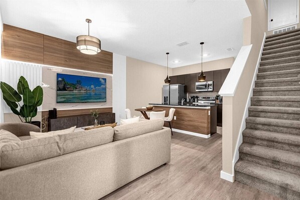 A modern living room with a sofa, a dining area, a kitchen, and a staircase.