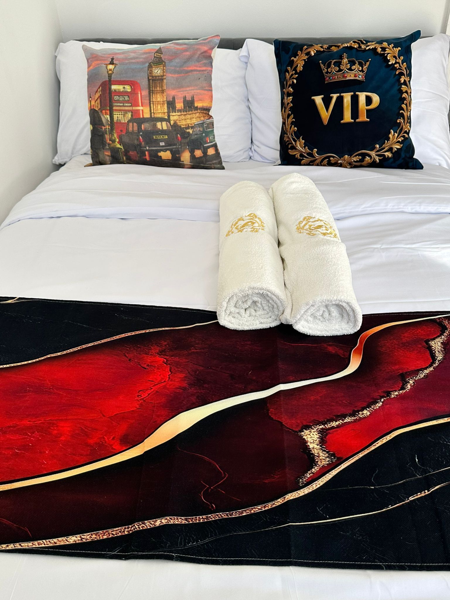 A neatly made bed with white linens, a red and black decorative throw, and two white towels with gold embroidery.