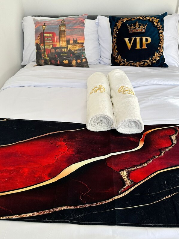 A neatly made bed with white linens, a red and black decorative throw, and two white towels with gold embroidery.