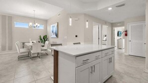 Una cocina moderna con armarios blancos, una isla central, iluminación colgante y una zona de comedor con mesa y sillas blancas.