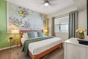 Un dormitorio con cama, mesita de noche, lámpara, florero con flores, ventana y un mural en la pared.
