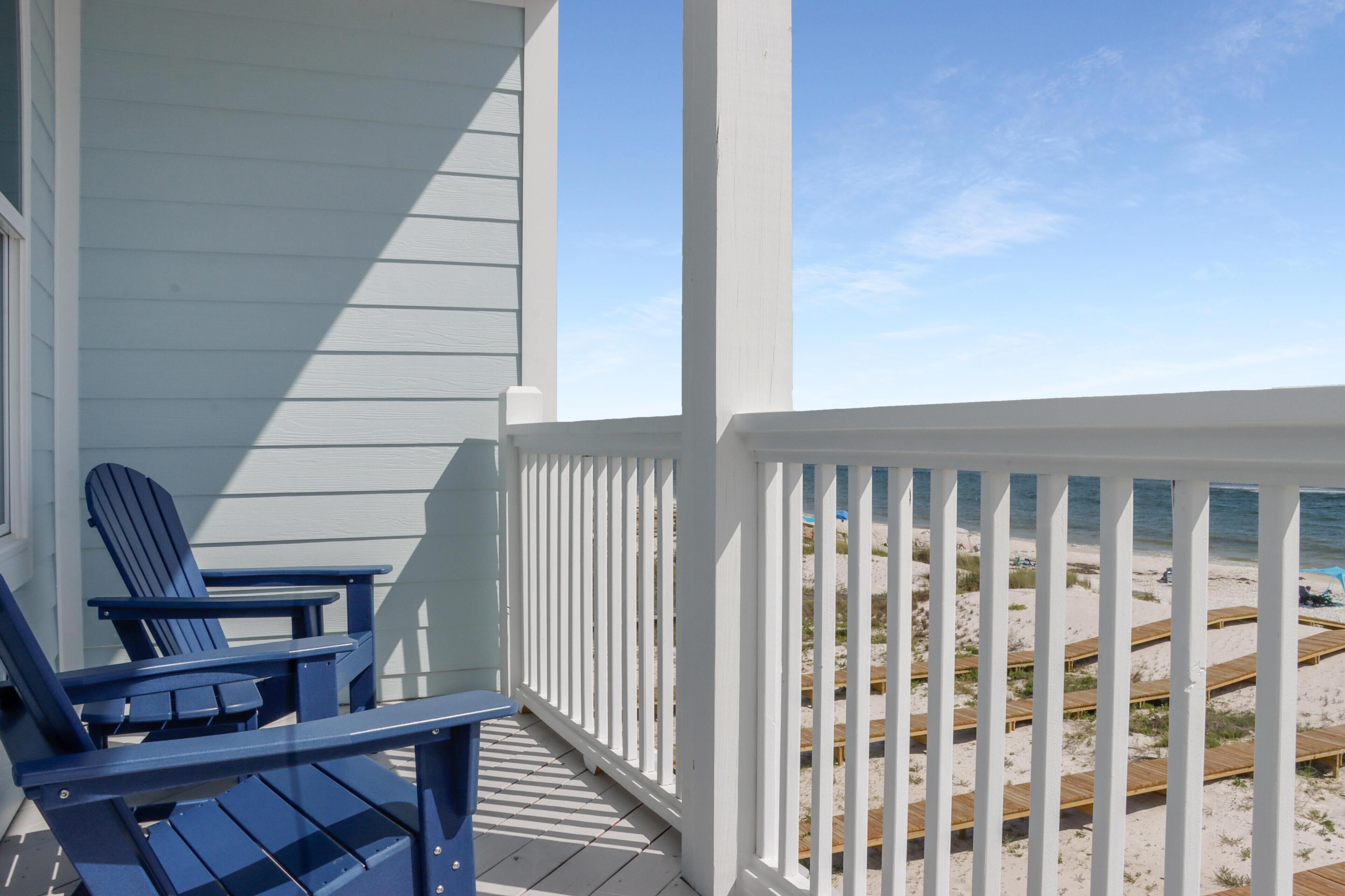 Una terraza de madera con dos sillas Adirondack azules, una baranda blanca y vista a una playa de arena con pasarelas de madera.