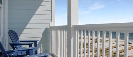 Una terraza de madera con dos sillas Adirondack azules, una baranda blanca y vista a una playa de arena con pasarelas de madera.
