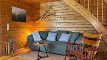 A cozy cabin interior with a wooden ceiling, a sofa, a rocking chair, a small table, and a mounted picture.