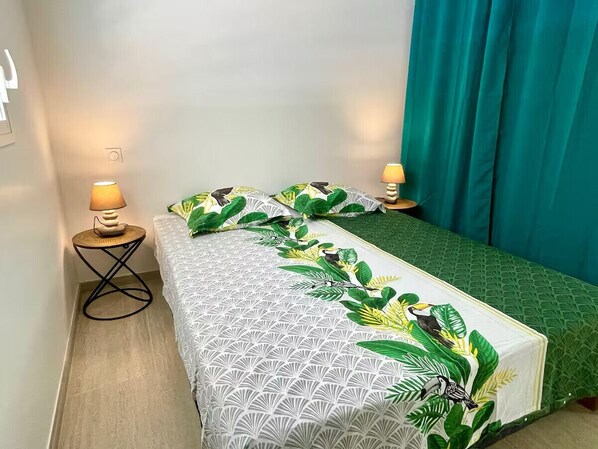 Una cama individual con una colcha de estampado geométrico y falda de cama verde con diseños de hojas tropicales. Hay dos lámparas de noche a cada lado de la cama.