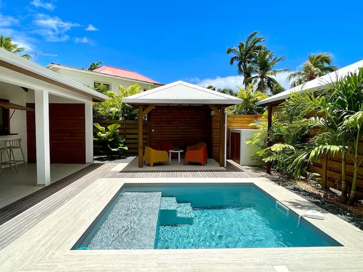 Une piscine avec une terrasse en bois, un espace salon couvert agrémenté de coussins et une vue sur un bâtiment au toit rouge.