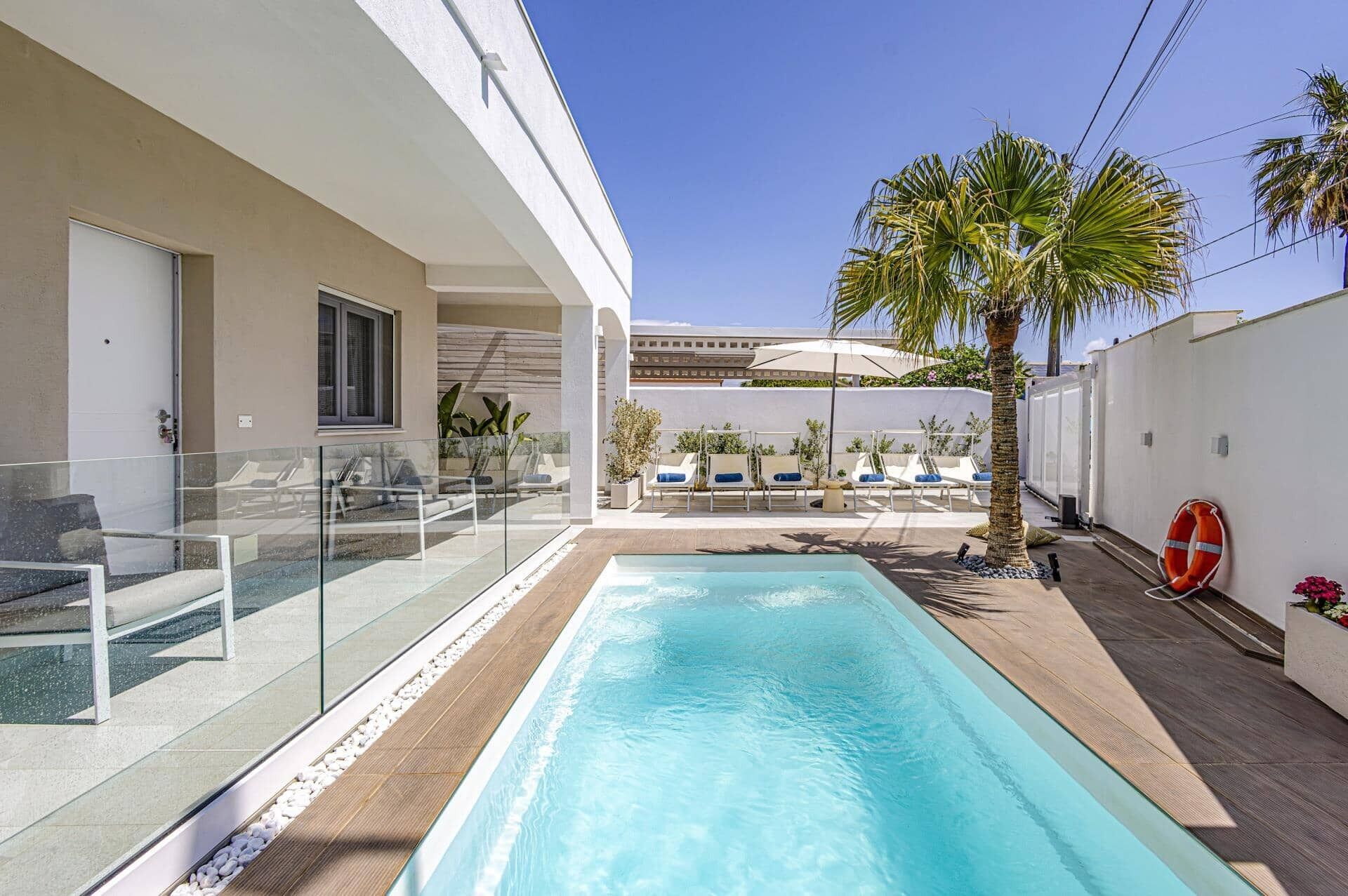 Un espace piscine moderne avec une eau d’un bleu limpide, entouré d’une rambarde en verre et d’une terrasse en bois. On aperçoit des chaises longues et un palmier à l’arrière-plan.