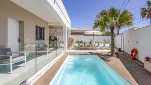 Un espace piscine moderne avec une eau d’un bleu limpide, entouré d’une rambarde en verre et d’une terrasse en bois. On aperçoit des chaises longues et un palmier à l’arrière-plan.