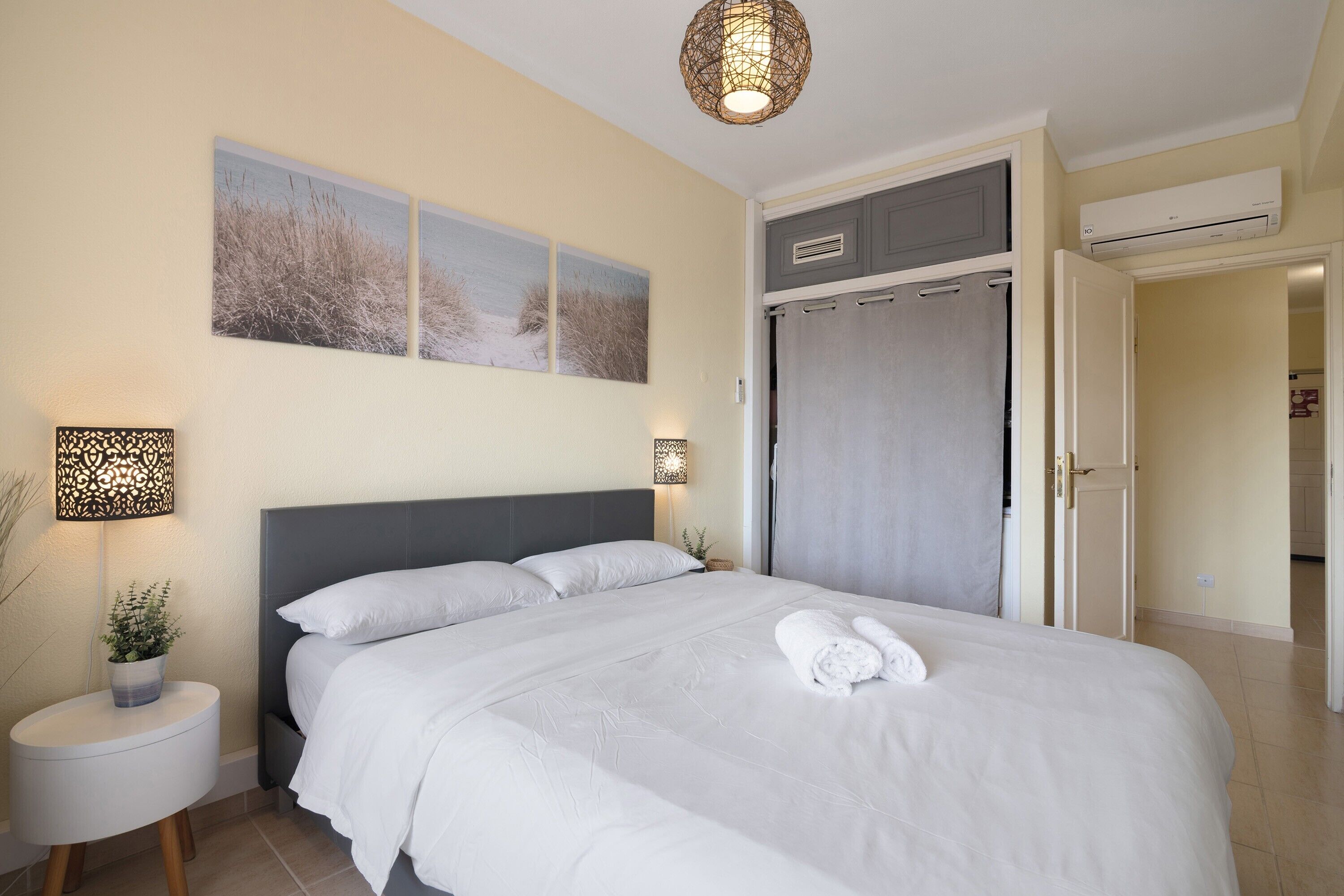 Ein ordentlich eingerichtetes Schlafzimmer mit einem Bett, Nachttischlampen, einer Wand-Klimaanlage und einer Tür zu einem anderen Raum.