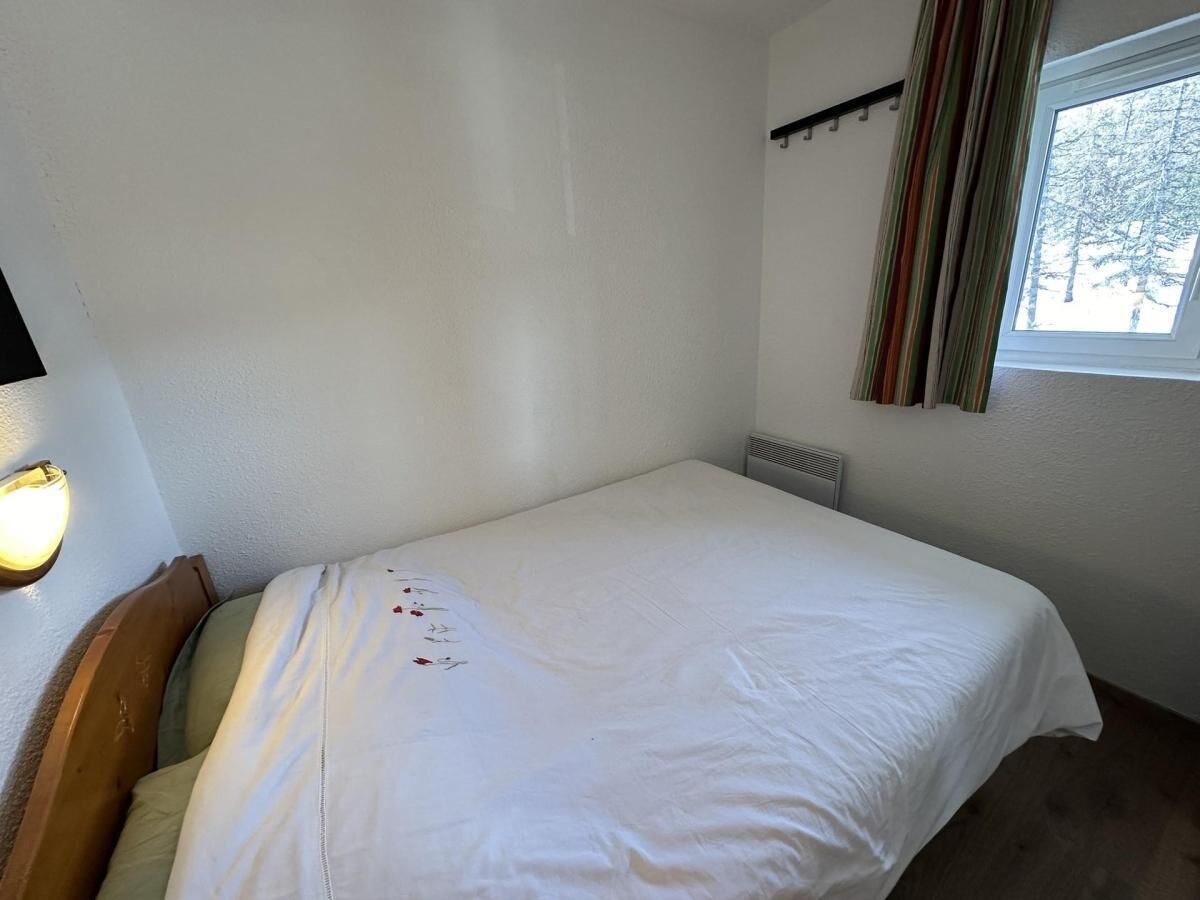 Une petite pièce simple, avec un lit simple, un radiateur, une fenêtre avec des rideaux et un luminaire fixé au mur.