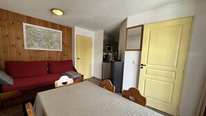 Una habitación con cama, un sofá rojo, una mesa de comedor con sillas, una zona de cocina y dos puertas.