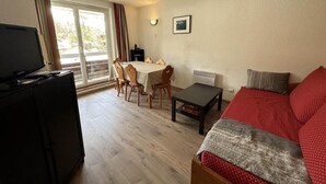 Una sala de estar con un sofá rojo, un televisor sobre un mueble, una mesa de comedor con sillas y una ventana con cortinas.